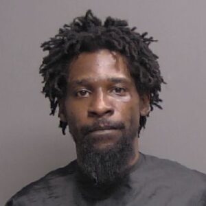 A man with short, twisted dreadlocks, a beard, and mustache, wearing a dark garment, poses against a plain gray background for a portrait style photograph