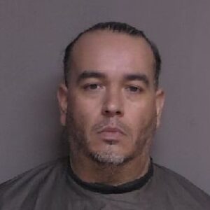 A middle aged man with short dark hair and a trimmed beard is facing the camera against a plain background, wearing a dark colored garment