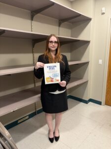 A woman in business attire stands in front of empty shelves, holding a paper that says "Falcon Fuel" with food images and logos on it