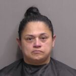 A woman with dark hair tied in a bun, wearing a black garment, is shown against a plain gray background, looking directly at the camera with a neutral expression