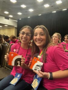 Two smiling young women in pink shirts sit side by side at an event, each holding a trophy They wear name tags and are surrounded by others sitting in rows of chairs in a large room