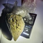 A clear plastic bag filled with cannabis buds sits on a digital scale, displaying a weight of grams The scene is lit with a flashlight, creating strong shadows and highlights on a white surface