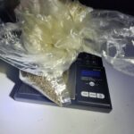 A plastic bag containing small brown seeds is placed on a digital weighing scale with a blue lit display showing numbers The scene is dimly lit, with shadows in the background