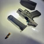 A black handgun with a magazine removed, two bullets, and a hard plastic gun case are displayed on a white surface under bright lighting