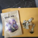 A brown packet labeled "Baby Ape" with colorful psychedelic mushroom artwork sits next to several dried mushrooms on a black marble surface