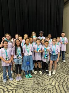 A group of children wearing light purple shirts and green medals stand together smiling in front of a black curtain, posing for a group photo on a patterned carpet