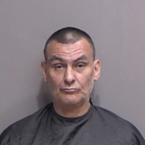 A middle aged man with short dark hair, wearing a black smock, stands against a plain gray background for a mugshot photo