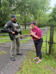 A law enforcement officer in tactical gear stands beside a metal gate, talking to a woman in a maroon shirt and black pants, who appears upset, with a grassy yard and trees in the background