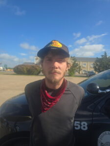 A young man with blond hair and a beard, wearing a black "Bass Pro Shops" cap, a red bandana, and a dark long sleeve shirt, stands in front of a police car on a sunny day