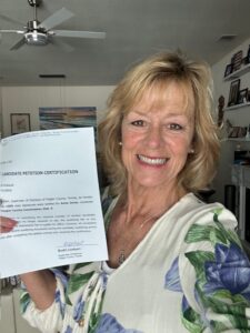 A smiling woman with blonde hair wearing a white floral blouse holds up a signed candidate petition certification in a well lit living room with art and shelves in the background