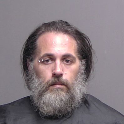 A man with long dark hair and a full gray beard looks at the camera He has a neutral expression, an eyebrow piercing, and is wearing a dark garment against a plain background