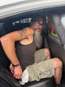 A man with a long gray beard, glasses, and a tank top sits handcuffed in the back seat of a police car, looking toward the camera