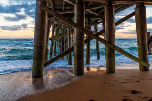 View from underneath a wooden pier at the beach, with ocean waves washing onto the sandy shore, footprints in the sand, and a cloudy sky at sunset in the background