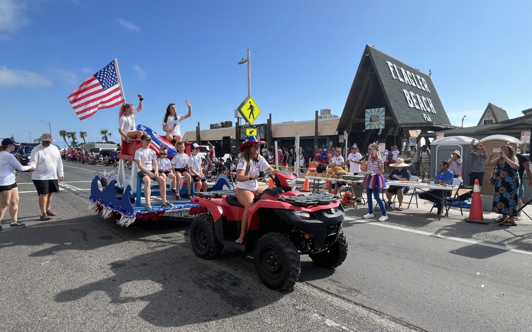A woman drives an ATV pulling a float with children in patriotic outfits and an American flag in a parade Spectators watch from the sidewalk near a building labeled "Flagler Beach