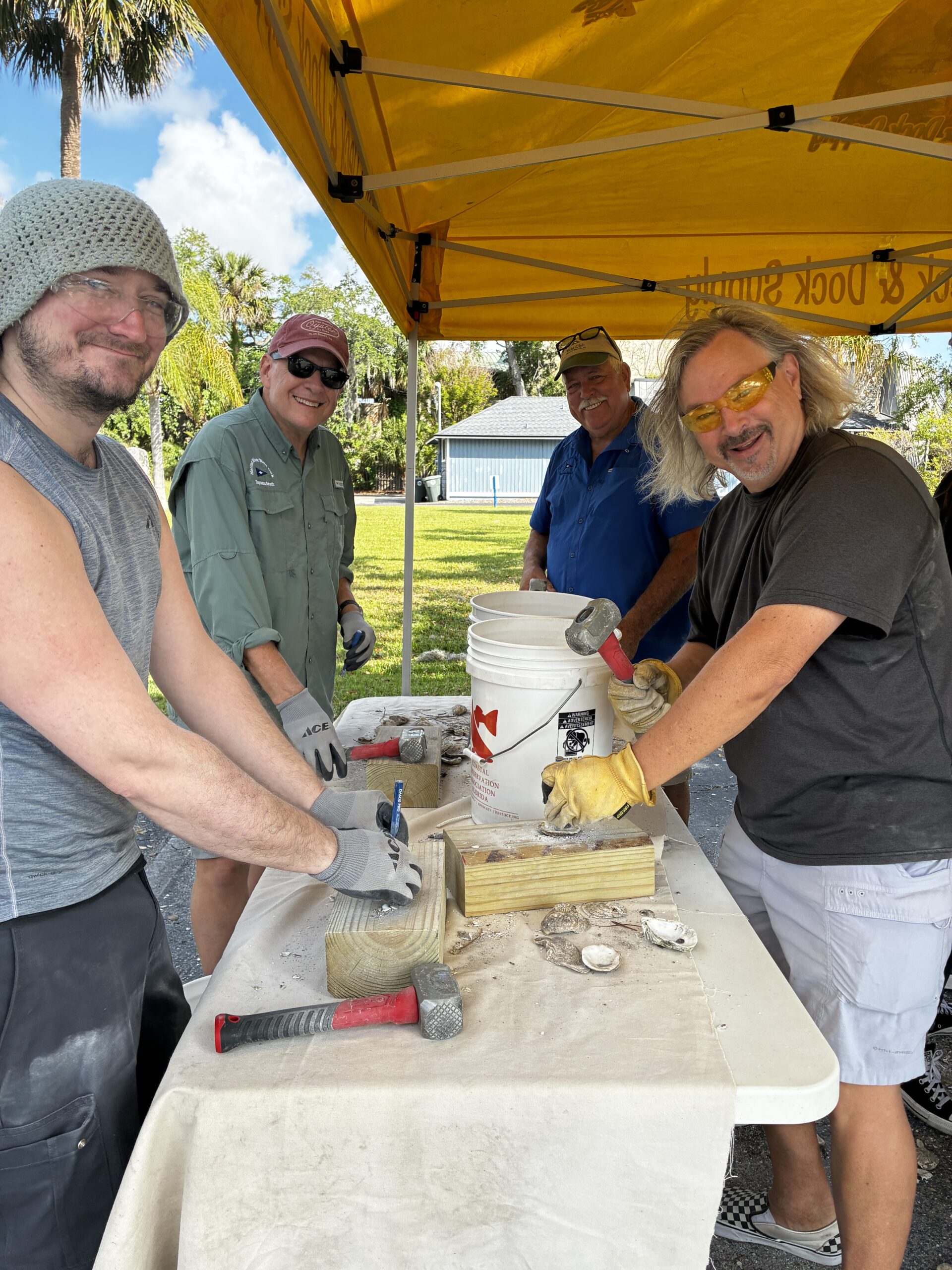 Four smiling men wearing protective gloves and goggles stand under a yellow canopy, working with hammers and molds at an outdoor table, surrounded by tools and a white bucket on a sunny day
