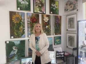 A smiling woman stands in front of a wall displaying colorful floral and nature themed paintings in a brightly lit art gallery