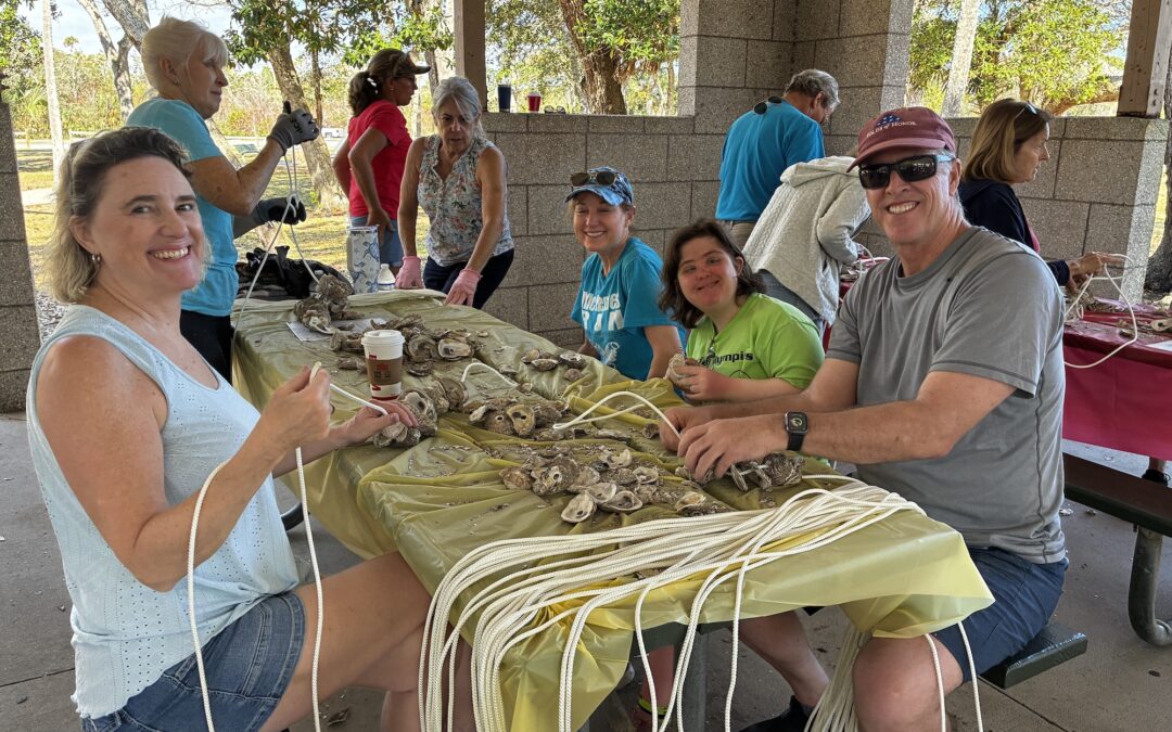 A group of smiling people sit around a table outdoors, stringing oyster shells together with white cords under a pavilion The table is covered with yellow cloth and various supplies Trees and more people are visible in the background