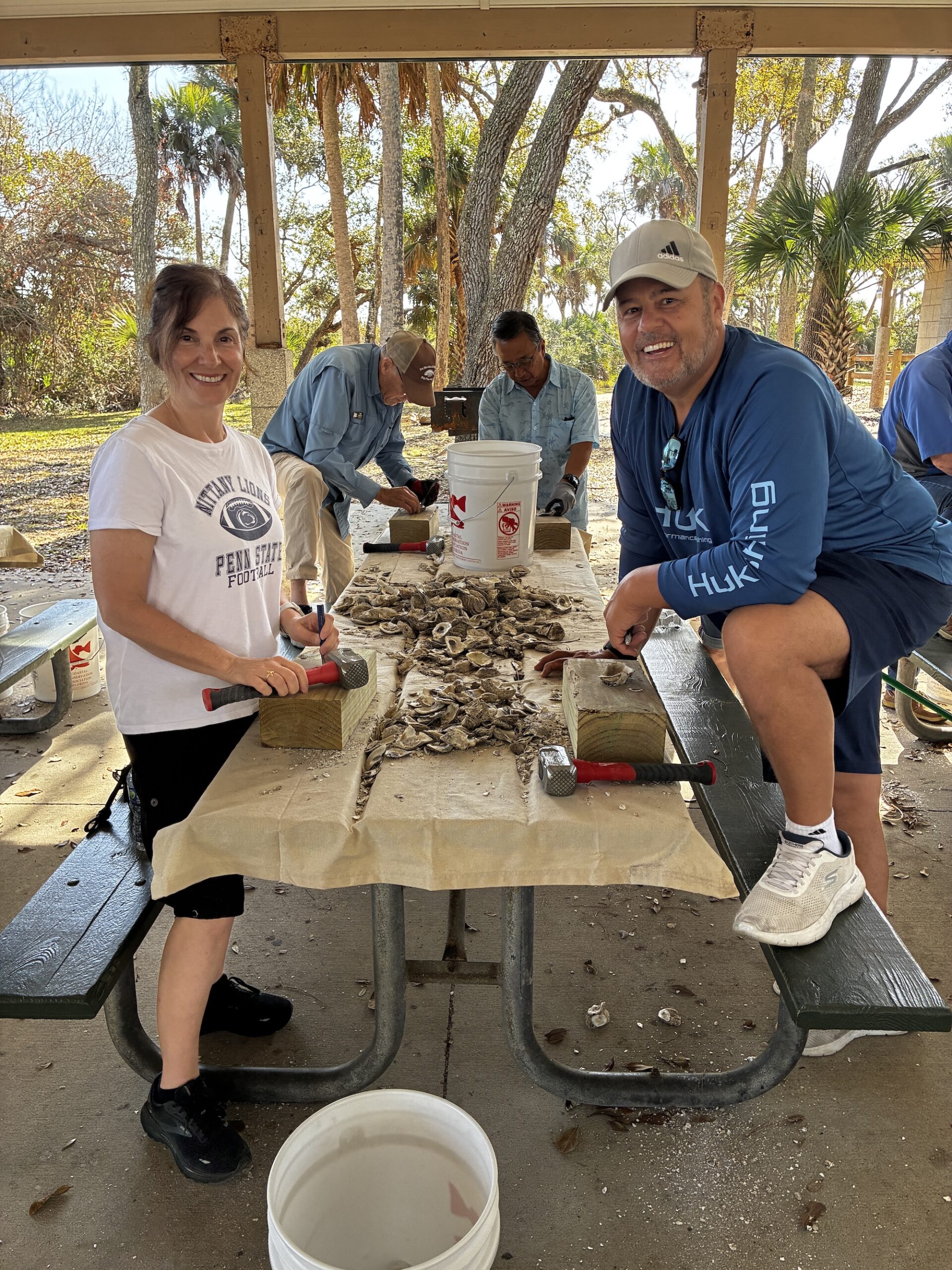 Two people smile at the camera while working on a table covered with oyster shells under a pavilion Other people in the background also work with oysters Buckets and tools are nearby, and trees can be seen outside