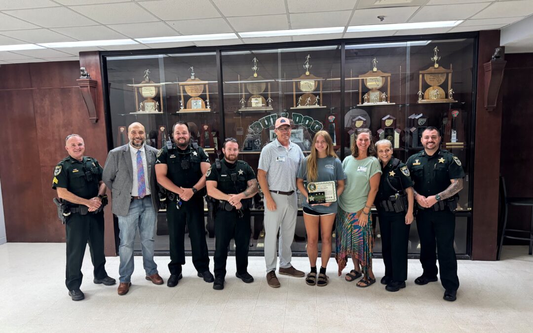 A group of law enforcement officers and civilians pose in front of a trophy display case A young woman holds an award