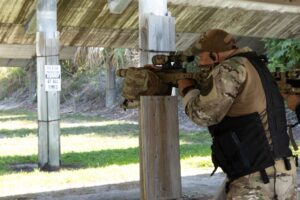 A person in camouflage and tactical gear aims a rifle while standing under a covered outdoor area A sign nearby reads, "Eye & Ear Protection Required At All Times