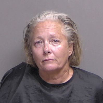 An older woman with gray hair wearing a black garment looks at the camera with a neutral expression against a plain background
