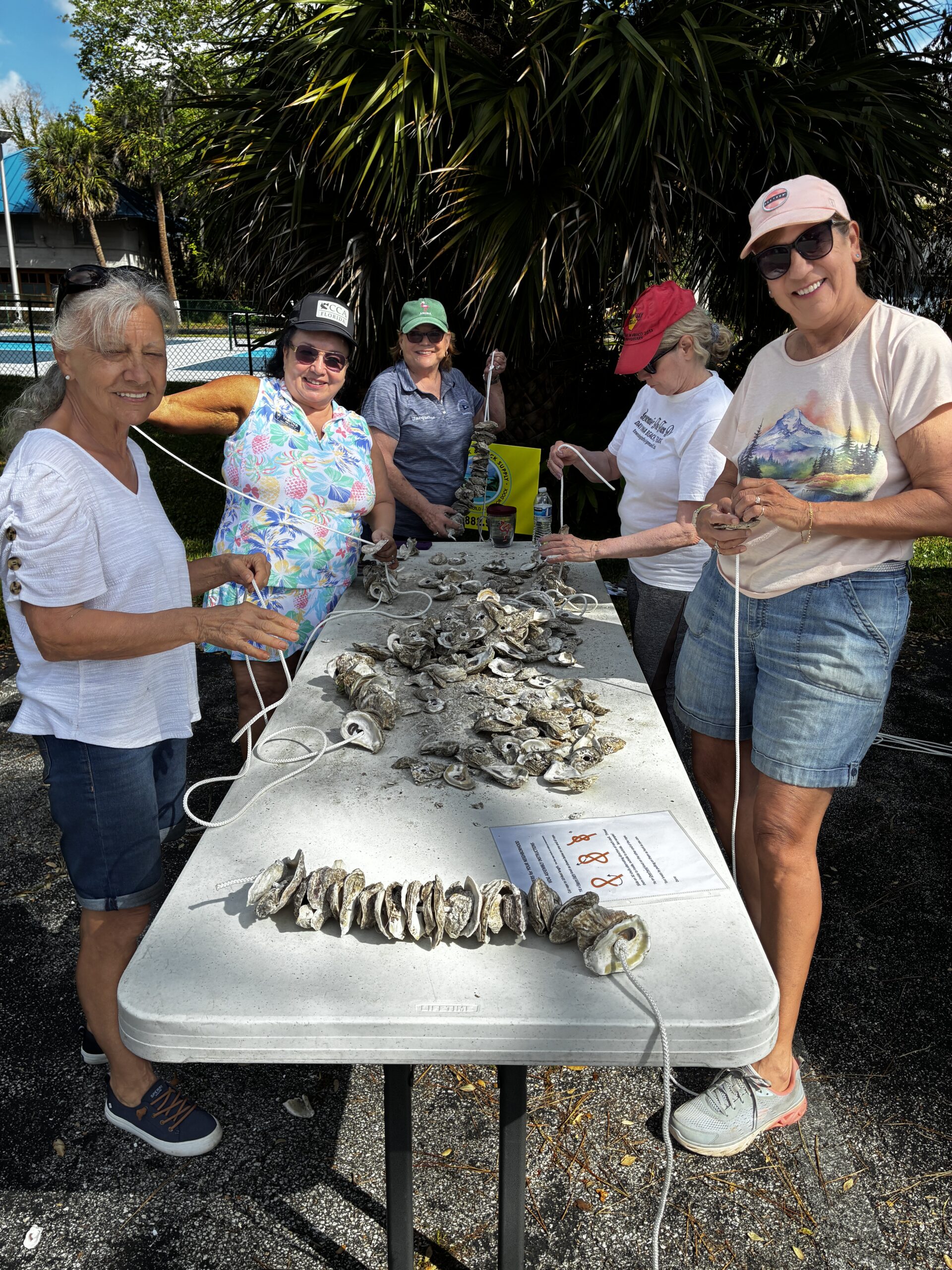 Five women stand around an outdoor table, threading oyster shells onto strings The table holds piles of shells, tools, and a paper with writing Palm trees and a fence are visible in the background on a sunny day