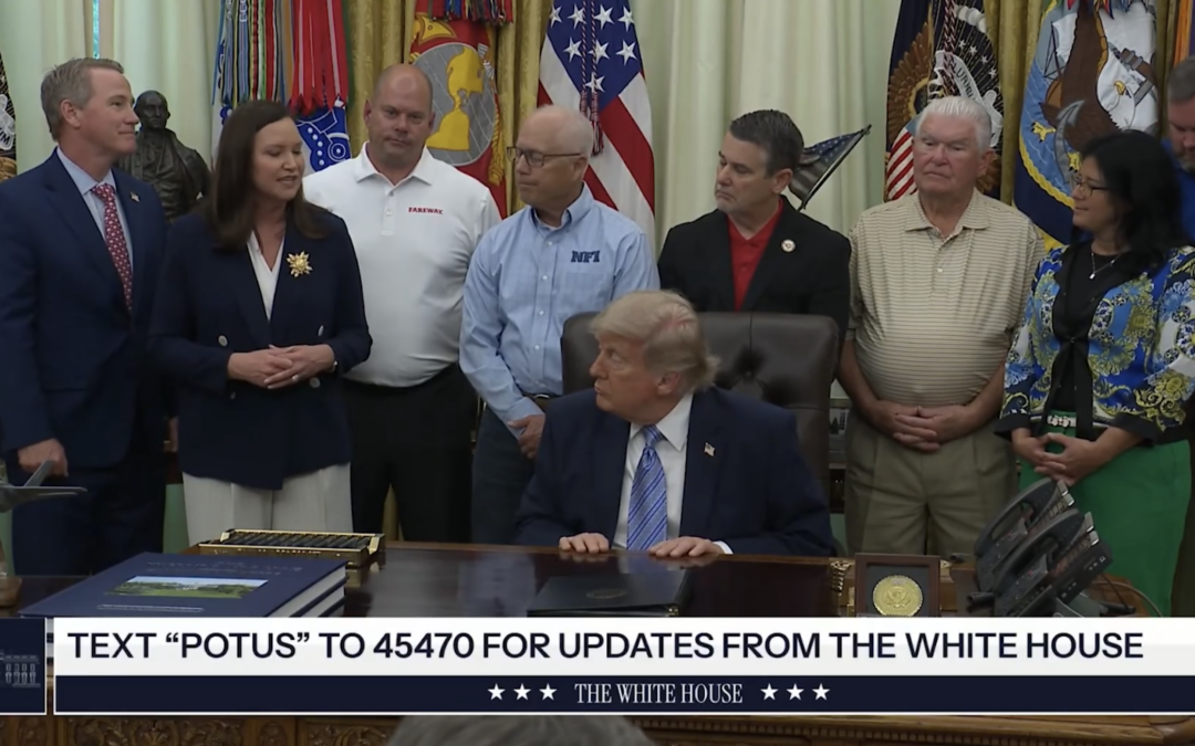 President Donald Trump sits at the Oval Office desk, looking at a group of people standing behind him Various U S and military flags are displayed in the background A text message update banner is visible at the bottom