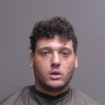 A young man with short, curly dark hair and facial stubble is facing the camera He is wearing a dark cape, as seen in mugshots, against a plain gray background