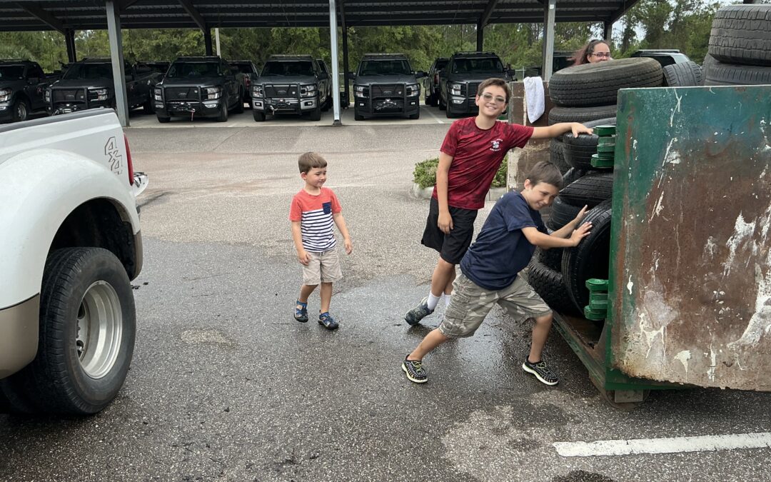 Three boys stand near a large container filled with tires in a parking lot; one boy pushes a tire, while the others watch Several black vehicles are parked under a carport in the background
