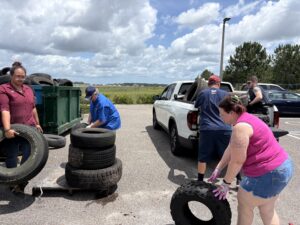 Four people load old tires into a green dumpster and a white pickup truck in a parking lot on a partly cloudy day, with grassy fields and trees in the background