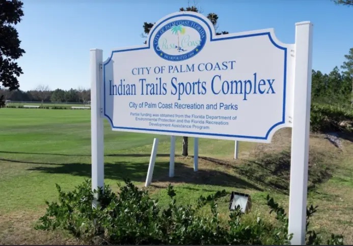 A white and blue sign reads “City of Palm Coast Indian Trails Sports Complex” with fields and trees in the background on a sunny day
