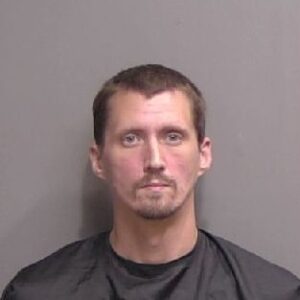 A man with short brown hair, light facial hair, and a neutral expression poses for a mugshot against a plain gray background He is wearing a dark drape commonly used in booking photos A man with short brown hair, light facial hair, and a neutral expression poses for a mugshot against a plain gray background He is wearing a dark drape commonly used in booking photos