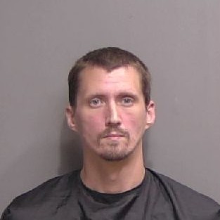 A man with short brown hair, light facial hair, and a neutral expression poses for a mugshot against a plain gray background He is wearing a dark drape commonly used in booking photos