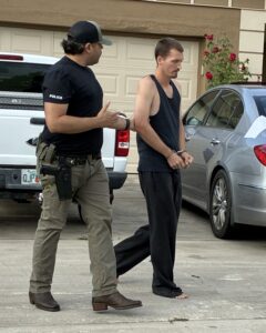 A police officer escorts a handcuffed man in casual clothes and no shoes on a residential driveway, with cars and a garage in the background A police officer escorts a handcuffed man in casual clothes and no shoes on a residential driveway, with cars and a garage in the background