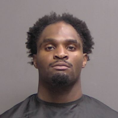 A man with short curly hair and a mustache looks straight at the camera in a mugshot style photo, wearing a dark garment against a plain gray background