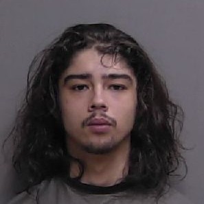 A young man with long dark hair and a mustache looks directly at the camera with a neutral expression, standing against a plain gray background