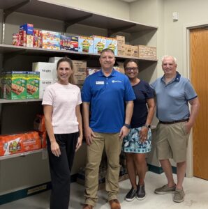 Four adults stand smiling in front of shelves stocked with snacks and drinks in a pantry or storage room The group is casually dressed and standing on a tile floor near a wooden door