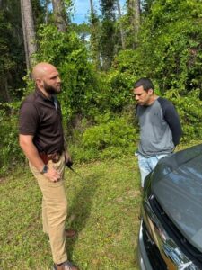 A bearded man in a brown shirt and tan pants, carrying a radio and holstered gun, stands beside a man in a gray shirt and jeans with his hands behind his back, near a vehicle and green trees A bearded man in a brown shirt and tan pants, carrying a radio and holstered gun, stands beside a man in a gray shirt and jeans with his hands behind his back, near a vehicle and green trees