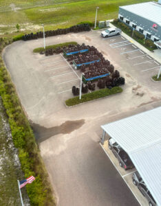 Aerial view of a parking lot with stacks of tires in multiple parking spaces, a few parked vehicles, an American flag at the lower left, and two buildings with metal roofs