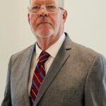 A man with short, light hair and glasses is wearing a gray suit, white shirt, and red and blue striped tie, standing against a plain light colored background A man with short, light hair and glasses is wearing a gray suit, white shirt, and red and blue striped tie, standing against a plain light colored background