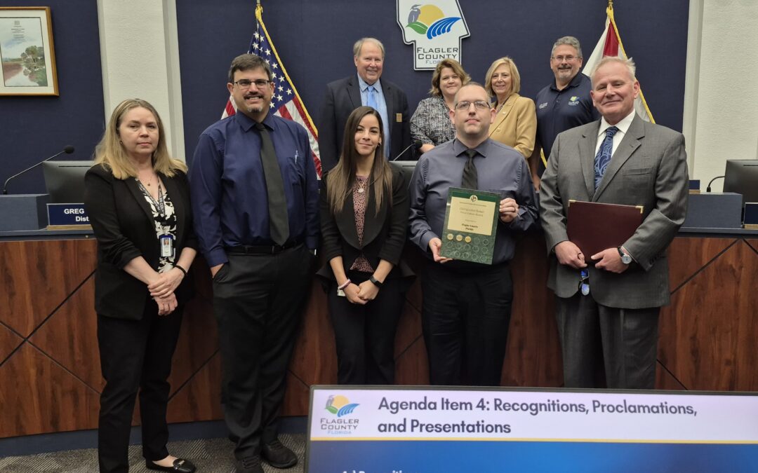 A group of nine people pose in a council chamber One person holds a plaque Behind them are two flags and a Flagler County seal A screen in the foreground displays an agenda for recognitions and proclamations