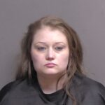 A woman with long brown hair and a neutral expression poses for a mugshot against a plain gray background, wearing a black drape