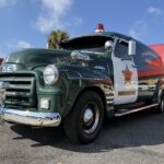 A vintage green GMC sheriff’s van with chrome details, parked in front of a building with a red roof on a sunny day