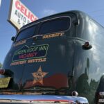 A vintage green sheriff's vehicle with "Green Roof Inn, Vacancy, Courtesy Shuttle" written on the back, parked near a sign reading "CELIC AUTO " Blue sky and trees are in the background
