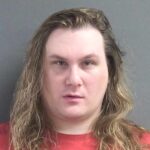 A person with long, wavy blond hair wears a red shirt and looks directly at the camera with a neutral expression The background is a plain gray wall