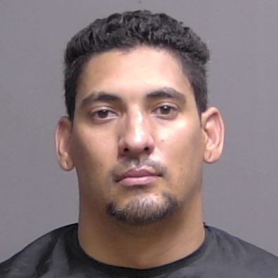 A man with short curly hair and a trimmed goatee is facing the camera with a neutral expression against a plain gray background