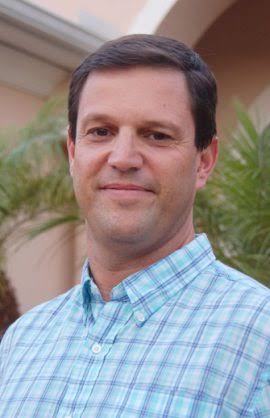 A man with short brown hair wearing a blue and white plaid shirt stands outdoors in front of palm trees and a peach colored building, smiling slightly at the camera