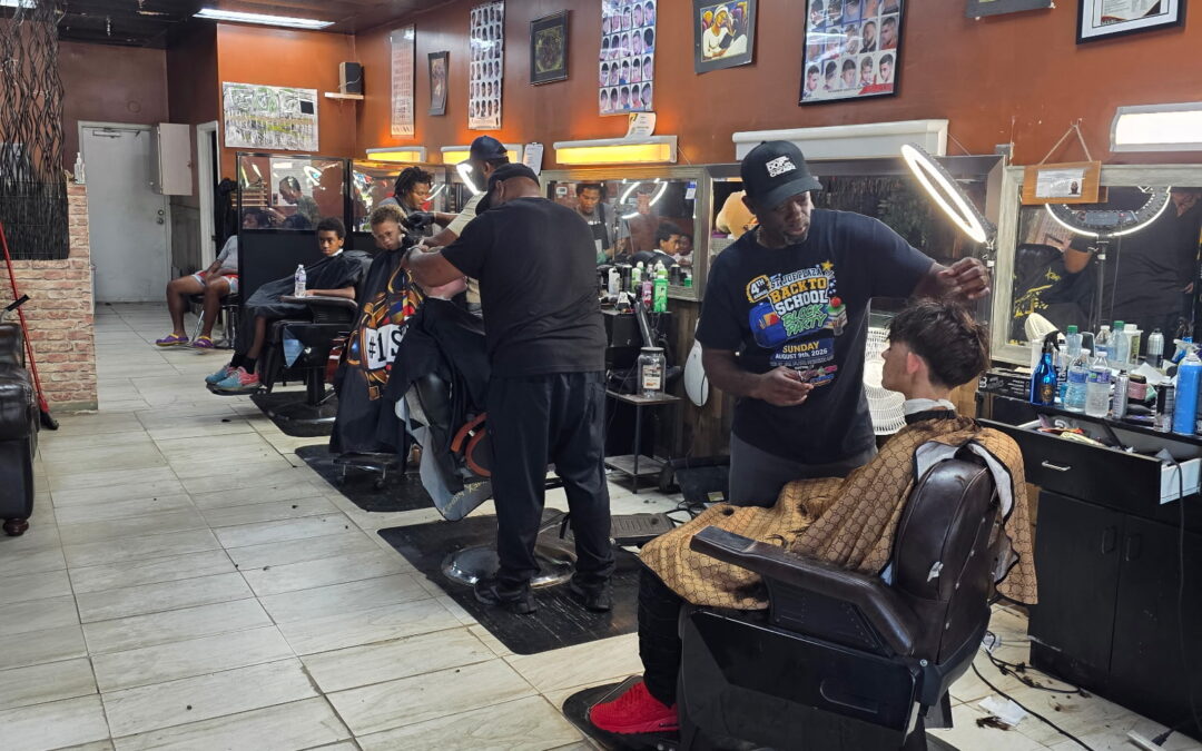 A busy barbershop with several barbers cutting hair, customers seated in barber chairs, and hair products on counters The walls display haircut style posters The shop has bright overhead lighting and tile floors