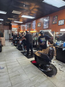 A busy barbershop with several barbers cutting hair, customers seated in barber chairs, and hair products on counters The walls display haircut style posters The shop has bright overhead lighting and tile floors
