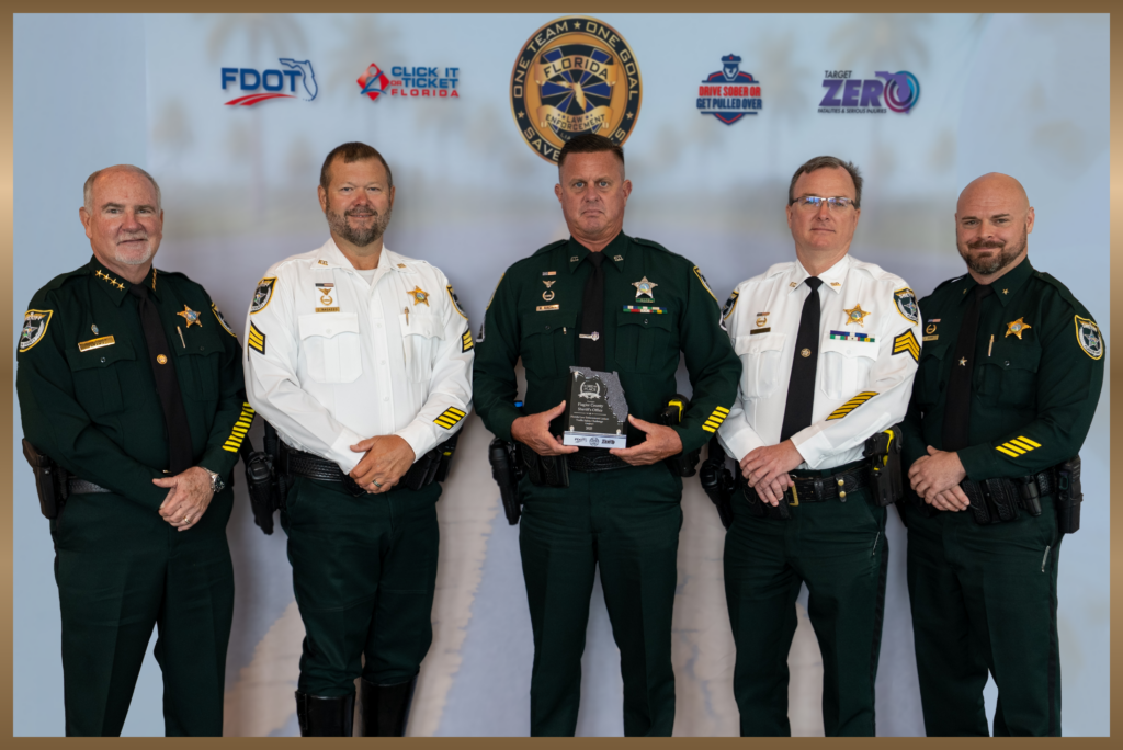 Five uniformed law enforcement officers stand indoors, with one holding a clear award Behind them is a backdrop featuring Florida safety campaign logos and a palm tree image Five uniformed law enforcement officers stand indoors, with one holding a clear award Behind them is a backdrop featuring Florida safety campaign logos and a palm tree image